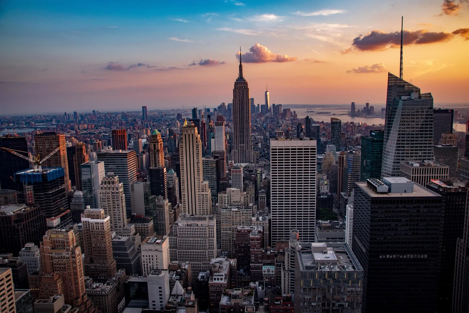 Midtown Manhattan from high above at sunset: the Empire State Building's Art Deco tower and spire rising in the centre of a dense field of skyscrapers, with One World Trade Center on the horizon behind it and the harbour beyond