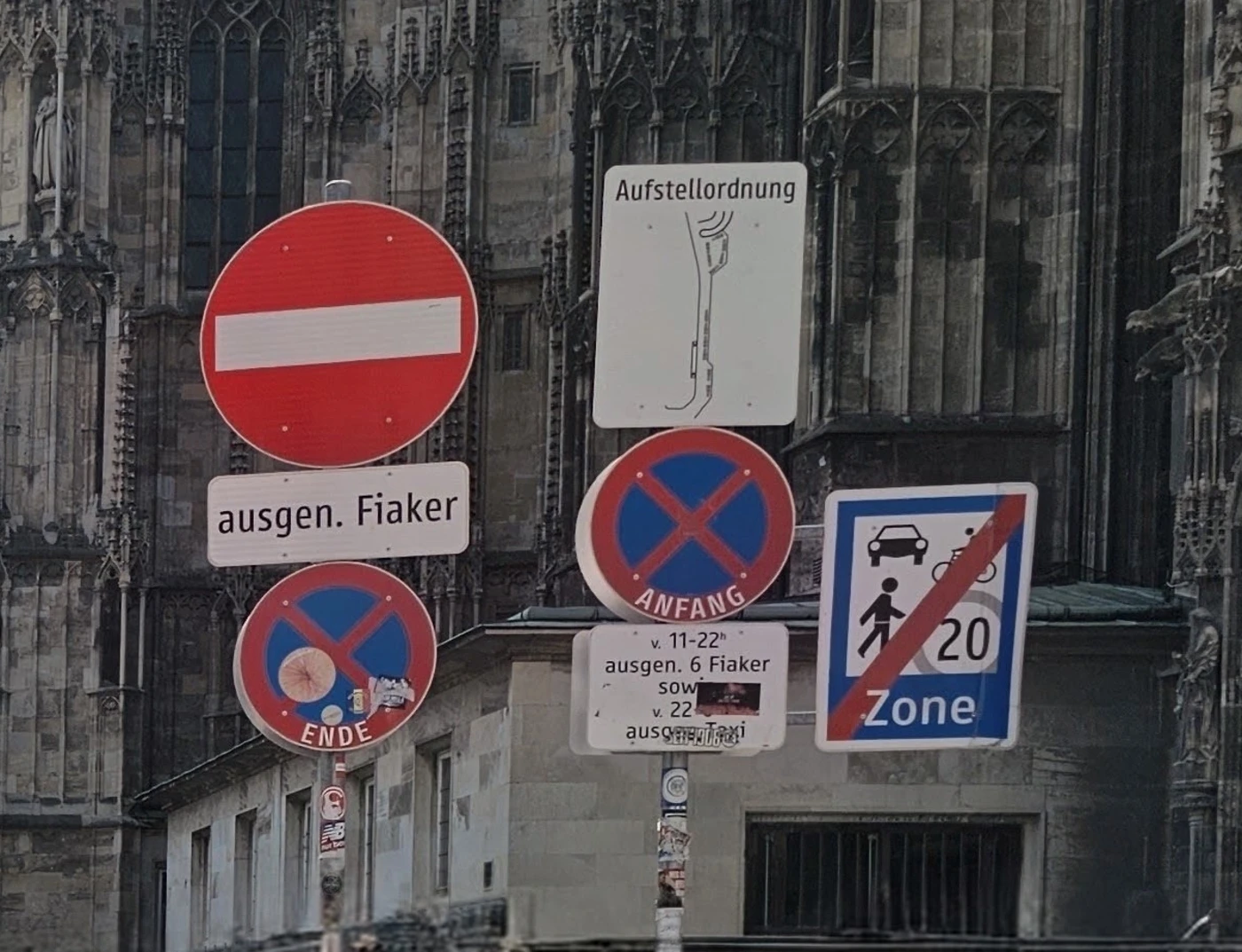 A cluster of Austrian street signs against Gothic cathedral stonework: a no-entry sign with an "ausgen. Fiaker" plaque, no-stopping signs with time restrictions, an Aufstellordnung sign, and an end-of-Begegnungszone sign with a 20 km/h limit