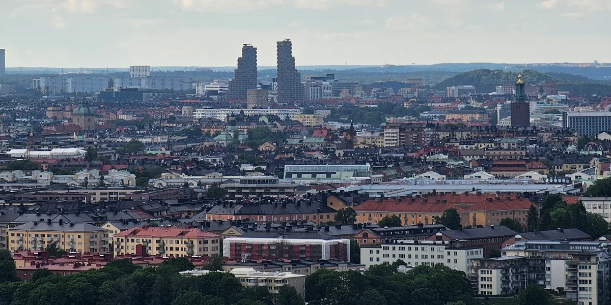 Stockholm skyline from the Avicii Arena SkyView on a hazy summer evening: a low spread of rooftops, Stockholm City Hall's tower with its three gilded crowns on the right, and the twin Norra Tornen towers rising in the middle