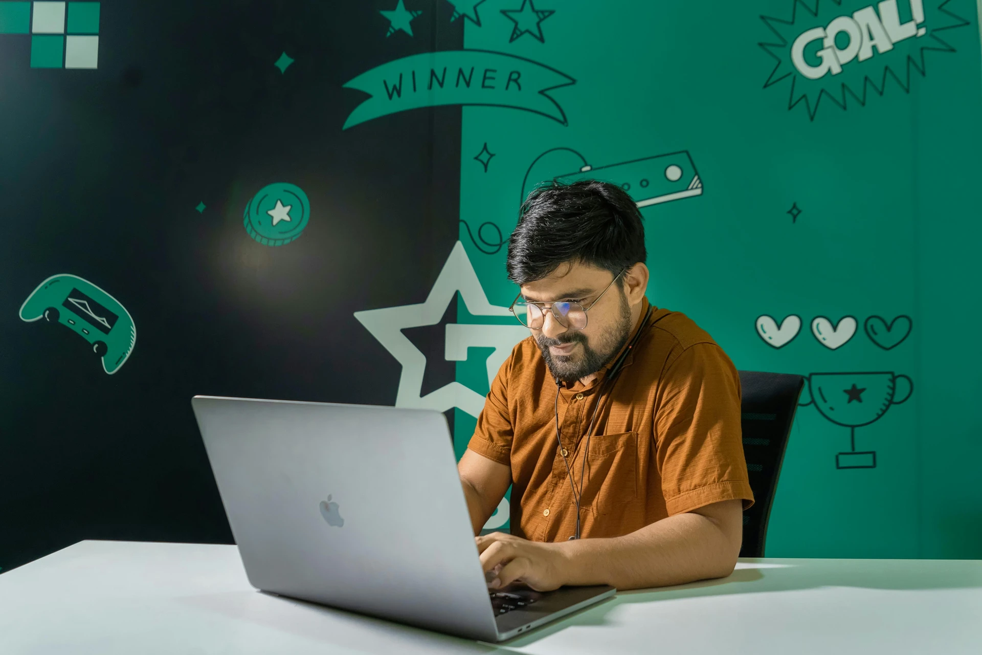 A man with a beard and glasses smiles while gaming on a laptop indoors, decorated with gaming graphics